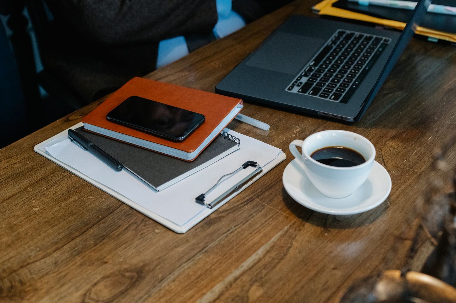 A phone resting on notebooks beside a laptop and a cup of coffee on a wooden desk